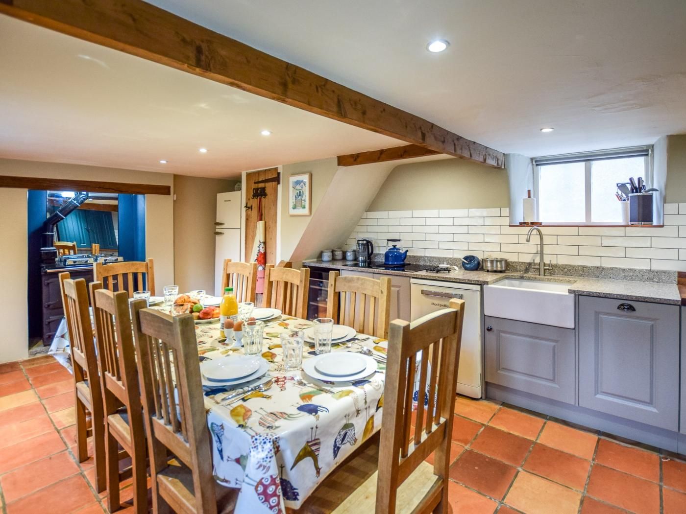 A dining area with a table set for four, wooden chairs, a kitchen with a sink and stove, and a window above the sink.