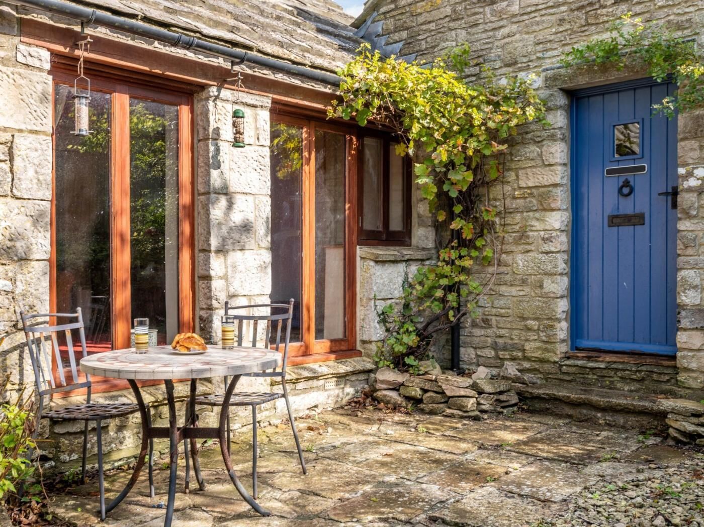 Un bâtiment en pierre avec une porte bleue, une table ronde entourée de chaises et une fenêtre ornée d’une lanterne suspendue.