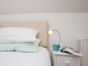 A neatly made bed with pillows, a bedside lamp, a small table with a mug and a magazine, and a radiator. - 3 Bed in Fowey (oc-pc008) (Fowey)