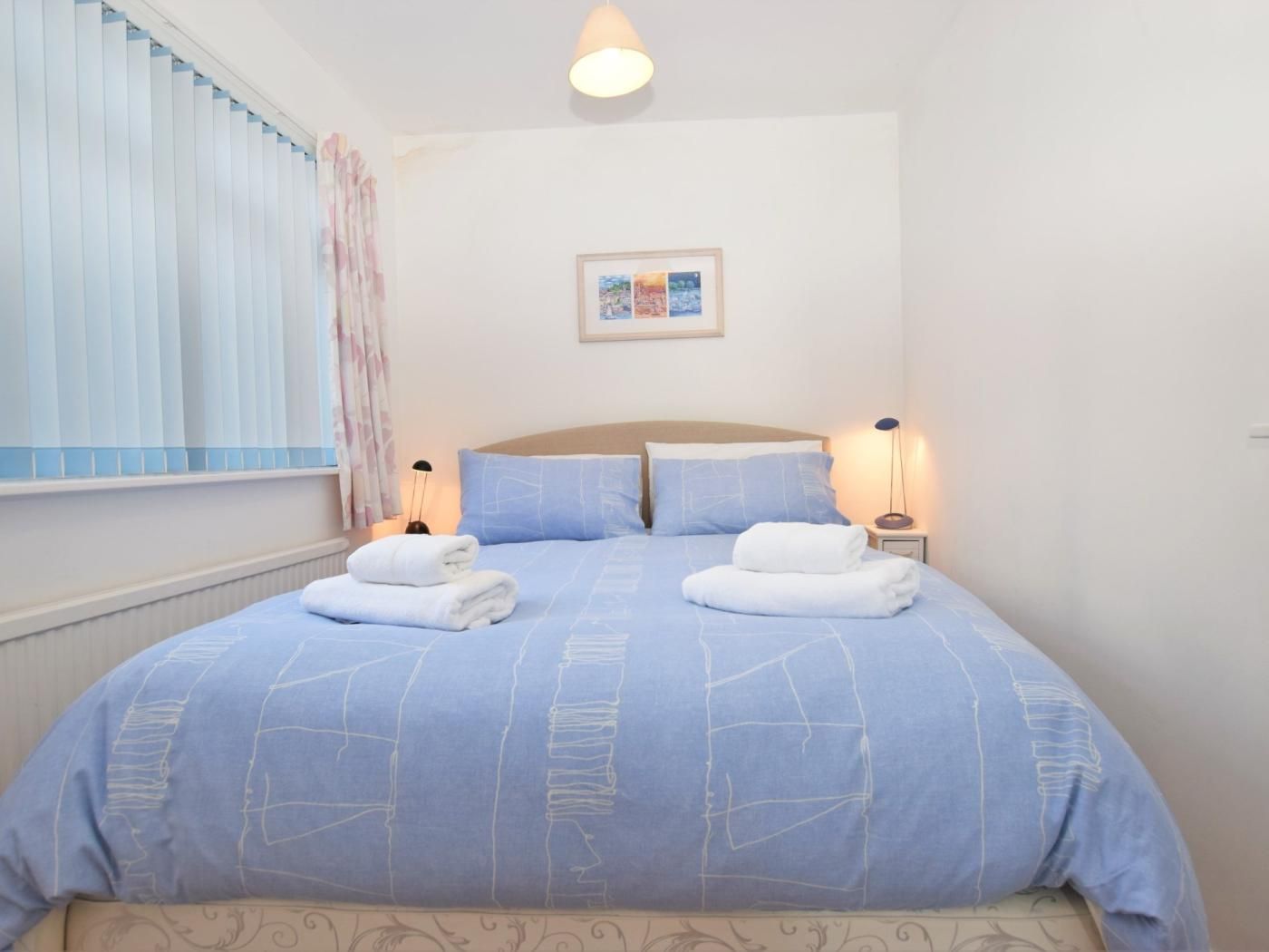A double bed with a blue bedspread and two pillows, a bedside table with a lamp, and a framed picture on the wall.