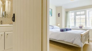 A bedroom with a bed, a window with curtains, a bedside lamp, and a bathroom with a mirror and sink. - 2 Bed in Polruan (oc-pc021) (Fowey)