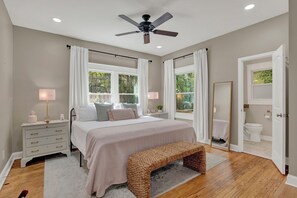 A bedroom with a bed, bedside tables, a bench, a ceiling fan, and a bathroom visible through an open door.
