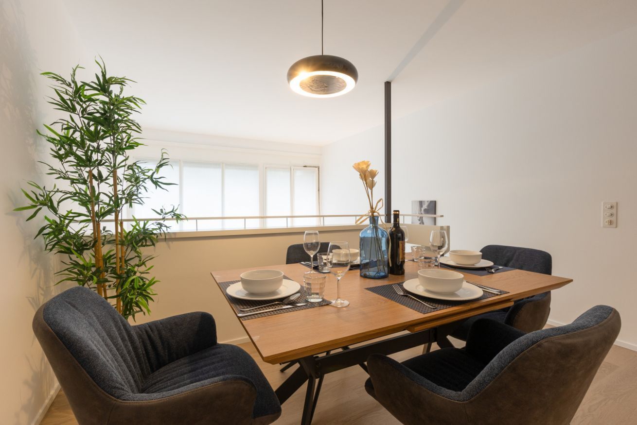 Un coin repas comprenant une table en bois, quatre chaises, une plante en pot, un vase avec des fleurs et un luminaire suspendu.