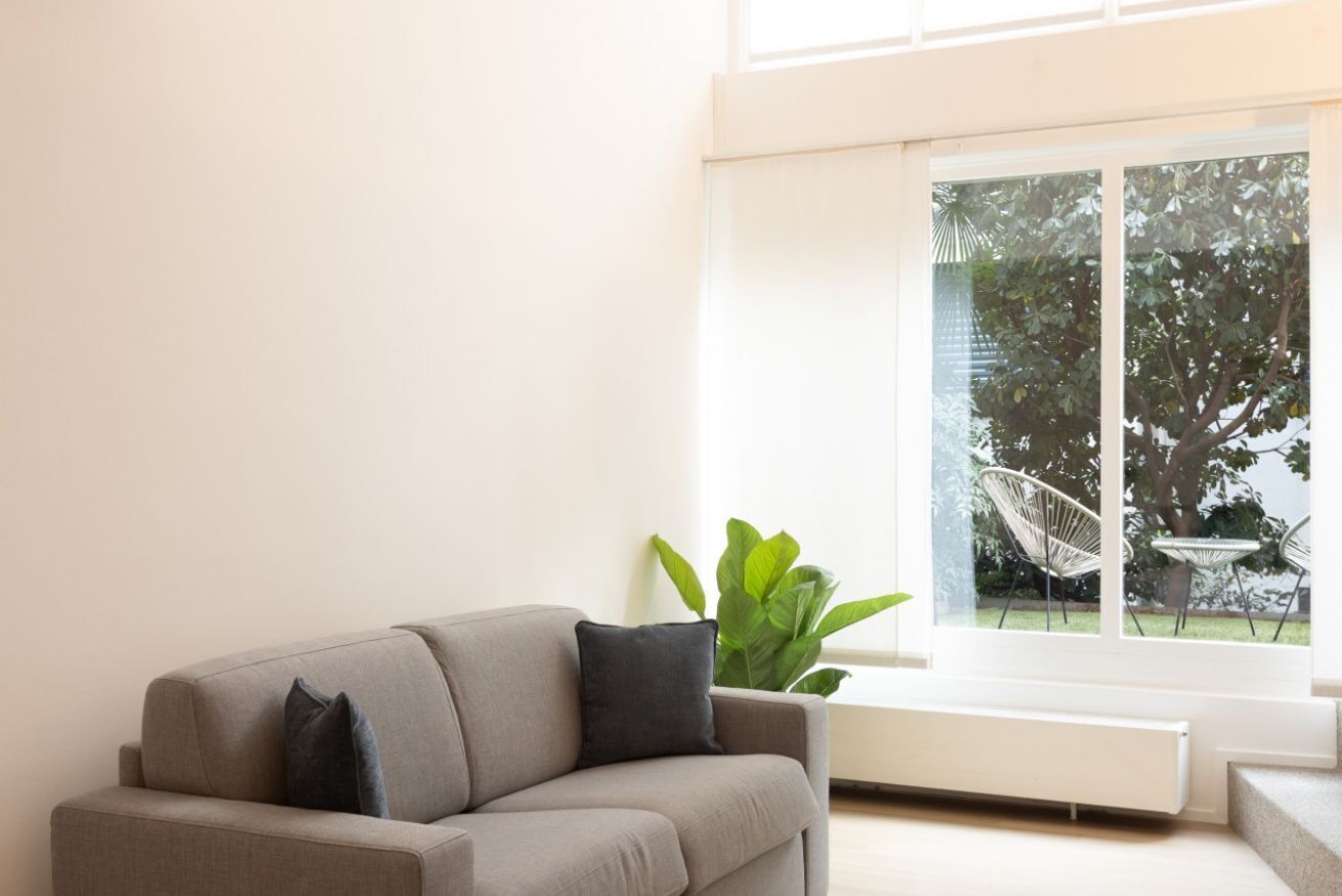Un salon moderne avec un canapé gris, une grande plante et une vue sur un jardin, visible à travers une grande fenêtre.