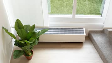 Una esquina de una habitación con una planta en maceta, una ventana que da a un área verde y un radiador blanco.