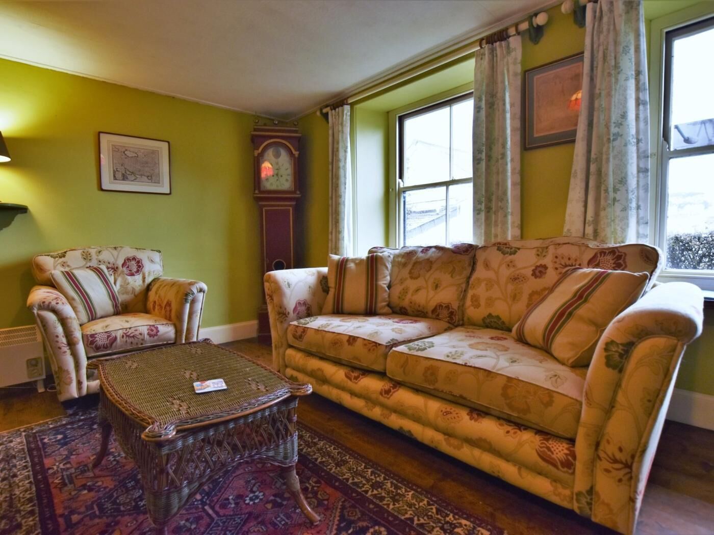 Een woonkamer met een bank, fauteuils, een rieten salontafel, een klok en een raam met gordijnen.