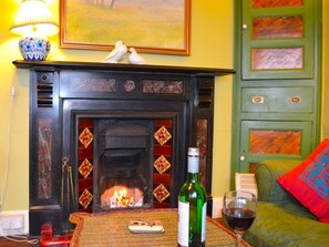 A fireplace with a fire, a table with a bottle and glass, and a green door with a colorful panel. - 3 Bed in Polruan (oc-pc032) (Fowey)