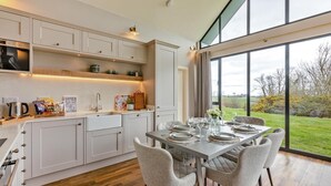 A modern kitchen with white cabinetry, a dining area with a table set for four, and a large window offering a view of the outdoors. - 3 Bed in Bamburgh (oc-61867) (Bamburgh)