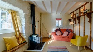 Ein gemütliches Zimmer mit Steinwand, einem schwarzen Ofen, einem rosafarbenen Sofa, einem gelben Sessel und einem Streifen Teppich.
