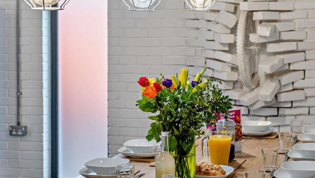 Mesa de comedor servida con platos, vasos, un jarrón con flores y una botella de jugo de naranja.