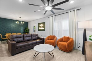 A modern living room with a brown leather sofa, two orange armchairs, a round coffee table, a ceiling fan, and a chandelier. - SA's Best Downtown Location - Terrace, Jazz Themed (San Antonio)