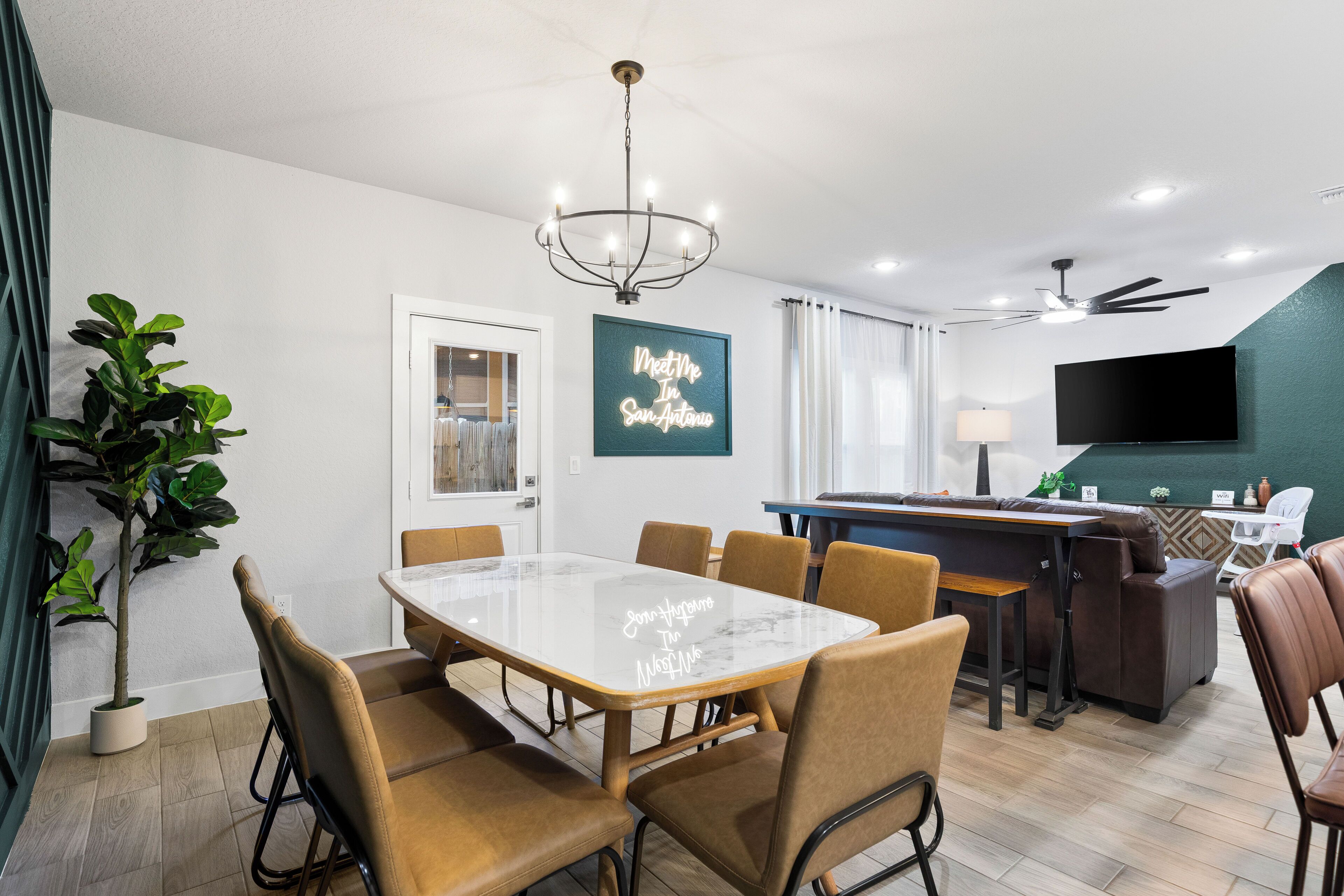 A modern dining area with a marble table, brown upholstered chairs, a chandelier, and a bar area with a television.