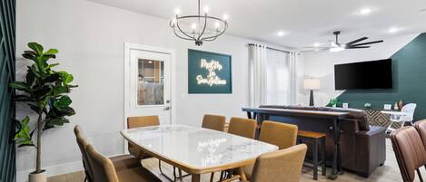 A modern dining area with a marble table, brown upholstered chairs, a chandelier, and a bar area with a television.