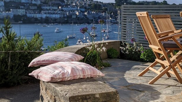 Een terras van natuursteen met twee kussens, een houten stoel en uitzicht op een haven met boten.