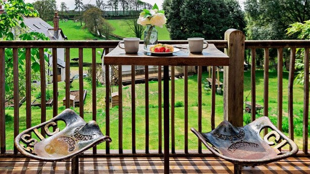 Ein Balkon mit einem für zwei Personen gedeckten Tisch, Blick auf eine grüne Landschaft und zwei alte Metallstühle.
