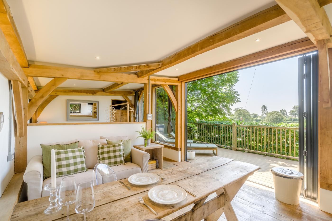 An open-plan living area with a wooden ceiling, a sofa set, a dining table with plates and glasses, and a view of greenery outside.