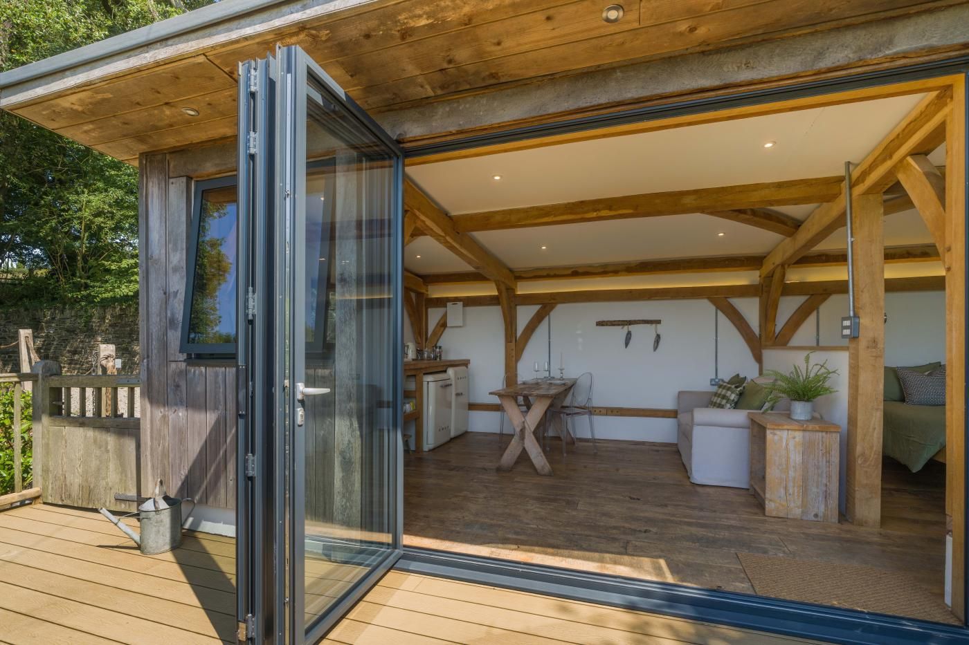 A modern wooden deck with a glass door leading to an interior space featuring a dining table, chairs, and a sofa.