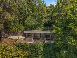 A modern house with a glass conservatory, surrounded by dense trees and a stone wall.