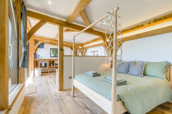 A bedroom with a four-poster bed, wooden beams, a small kitchenette, and a view of the outdoors.