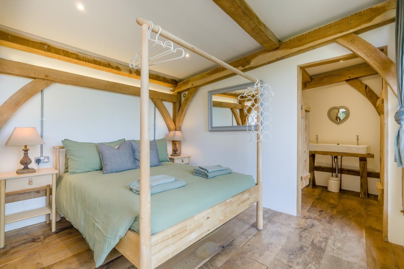 A bedroom with a four-poster bed, a bedside table with a lamp, a mirror, and a view of a bathroom with a sink and a heart-shaped mirror.