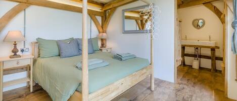 A bedroom with a four-poster bed, a bedside table with a lamp, a mirror, and a view of a bathroom with a sink and a heart-shaped mirror.