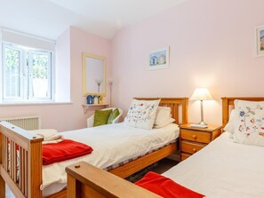 A bedroom with two beds, a window, a radiator, a bedside table with a lamp, and a small shelf with decorative items. - 3 Bed in Polruan (oc-pc012) (Polruan)