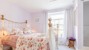 Een slaapkamer met een bed, een radiator, een raam met gordijnen en een klein tafeltje met een kussen.