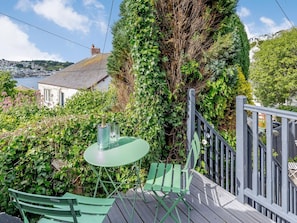 A green outdoor seating area with a table and chairs, surrounded by lush greenery and overlooking a coastal town.