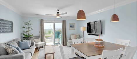A living room with a sofa, dining table, chairs, ceiling fan, pendant lights, TV, and a view through a glass door.