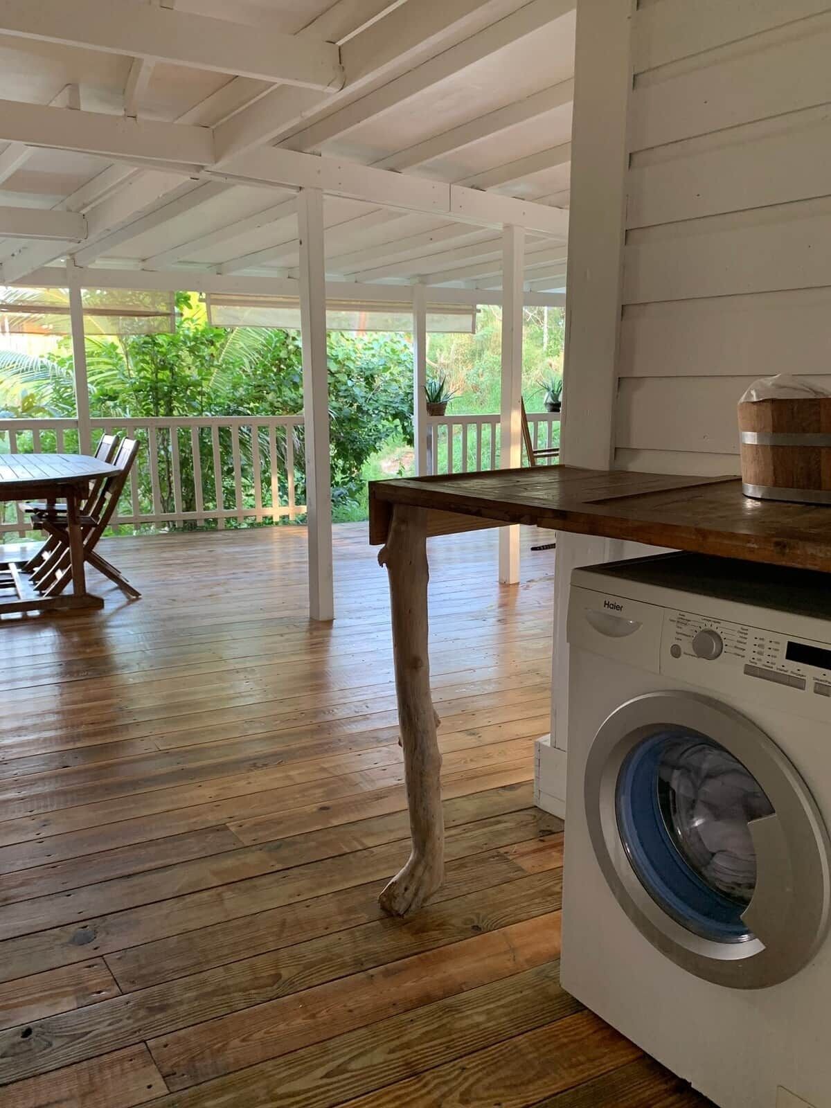 A covered porch with a wooden table and chairs, a washing machine, and a view of greenery.