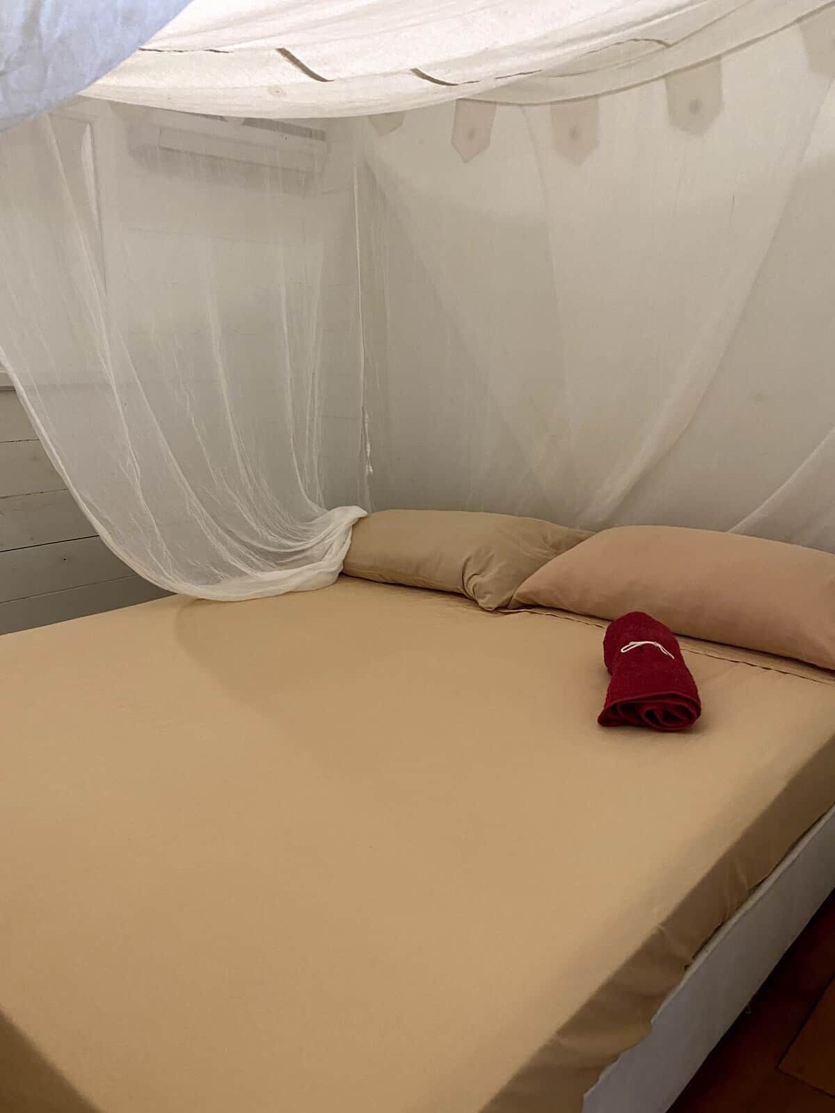 Una cama individual con un colchón color beige, una toalla roja y un mosquitero blanco.