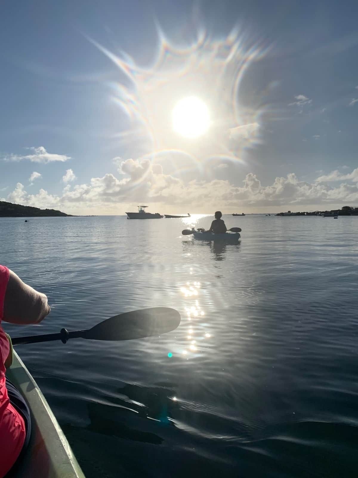 A calm body of water with a person paddling a kayak, a boat in the distance, and a clear sky.