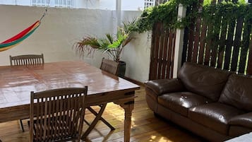 A wooden dining table with chairs, a brown leather sofa, a hammock, and potted plants in a covered outdoor area.