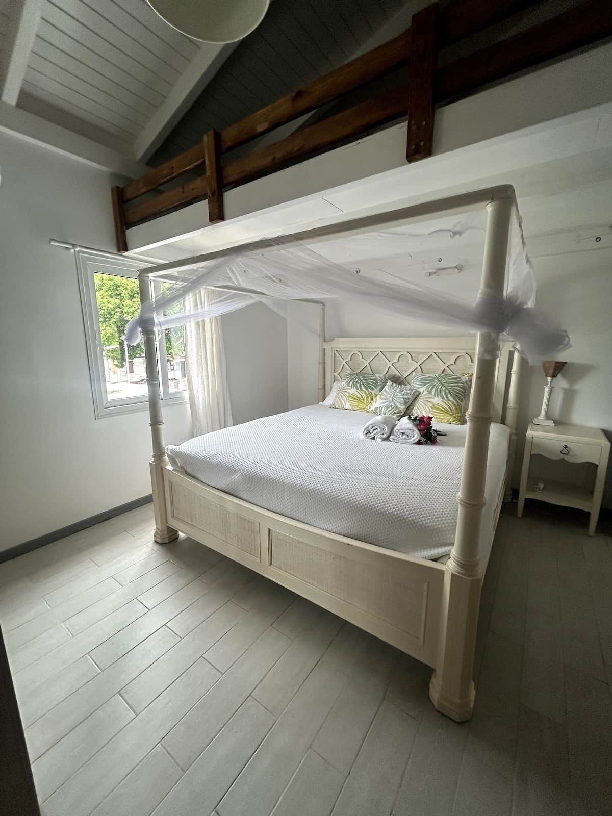 A four-poster bed with a canopy, a nightstand, and a window with a view of greenery.