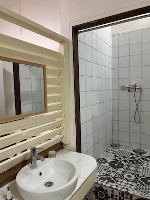 A bathroom with a white sink, a mirror with wooden blinds, and a shower area with patterned tiles. - Private Apartment in the Heart of Downtown Marigot (Marigot)