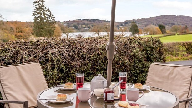 Une table ronde avec un plateau en verre, entourée de chaises de couleur beige, installée à l’extérieur et offrant une vue sur une rivière et des collines.