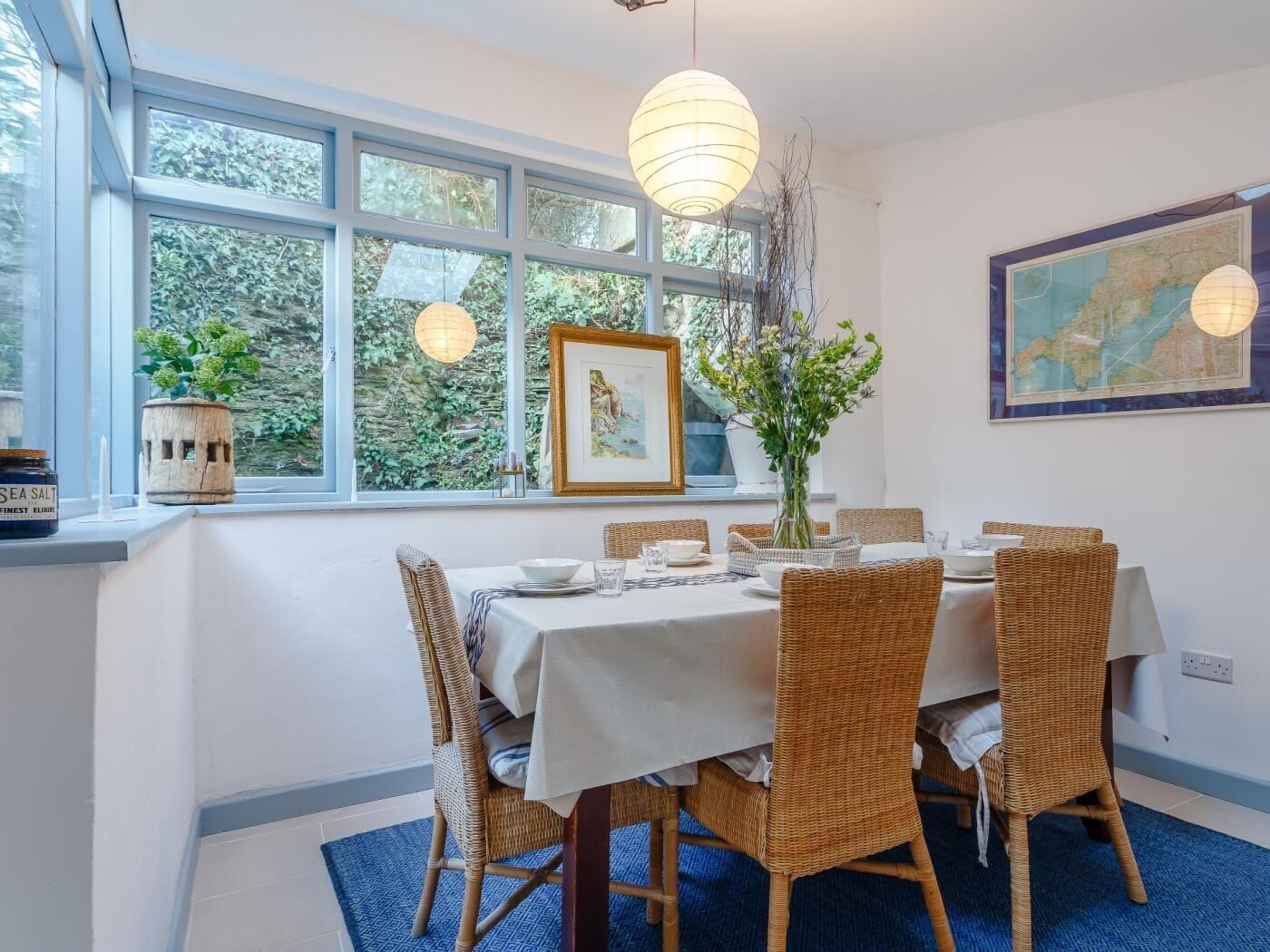 A dining area with a table set for four, wicker chairs, a vase with flowers, a framed picture, and a map on the wall.