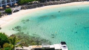 Une plage à l’eau turquoise et limpide, une jetée avec un bateau, et un hôtel en arrière-plan.