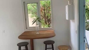 Een kamer met een houten tafel, twee zwarte barkrukken, een raam met uitzicht op groen, en een deur met een blauwe omlijsting.