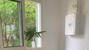 Une cuisine avec une table en bois, deux tabourets, une fenêtre donnant sur un espace verdoyant et un appareil électroménager blanc fixé au mur.