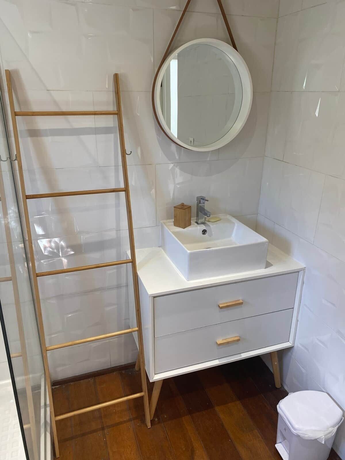 Ein Badezimmer mit weißem Waschtisch, einem runden Spiegel, einer Holzleiter und einer weißen Toilette.
