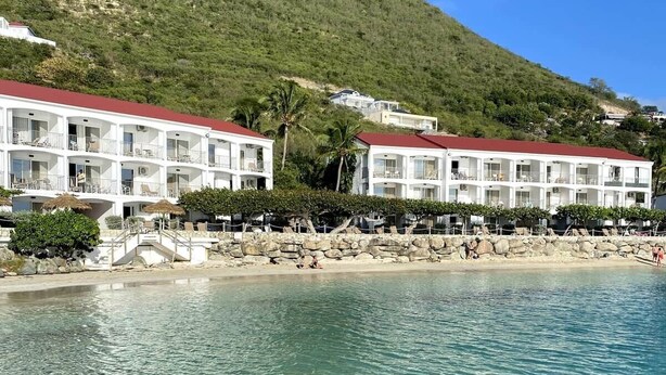Un hotel frente a la playa con aguas cristalinas, un edificio de techo rojo y un paisaje montañoso de fondo.