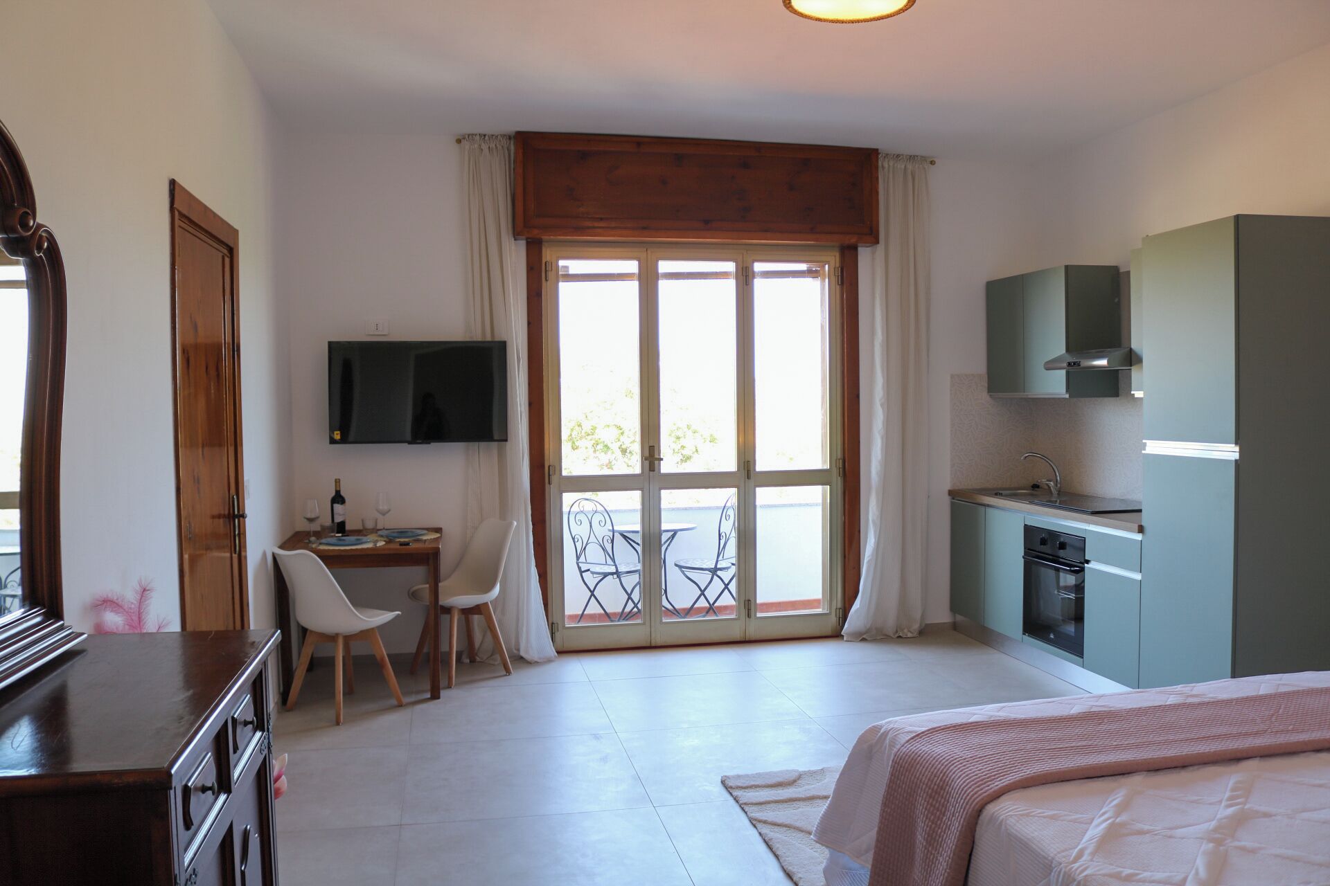 Una stanza con un letto, una scrivania con una sedia, una televisione, un angolo cottura e una porta-finestra che dà su uno spazio esterno.