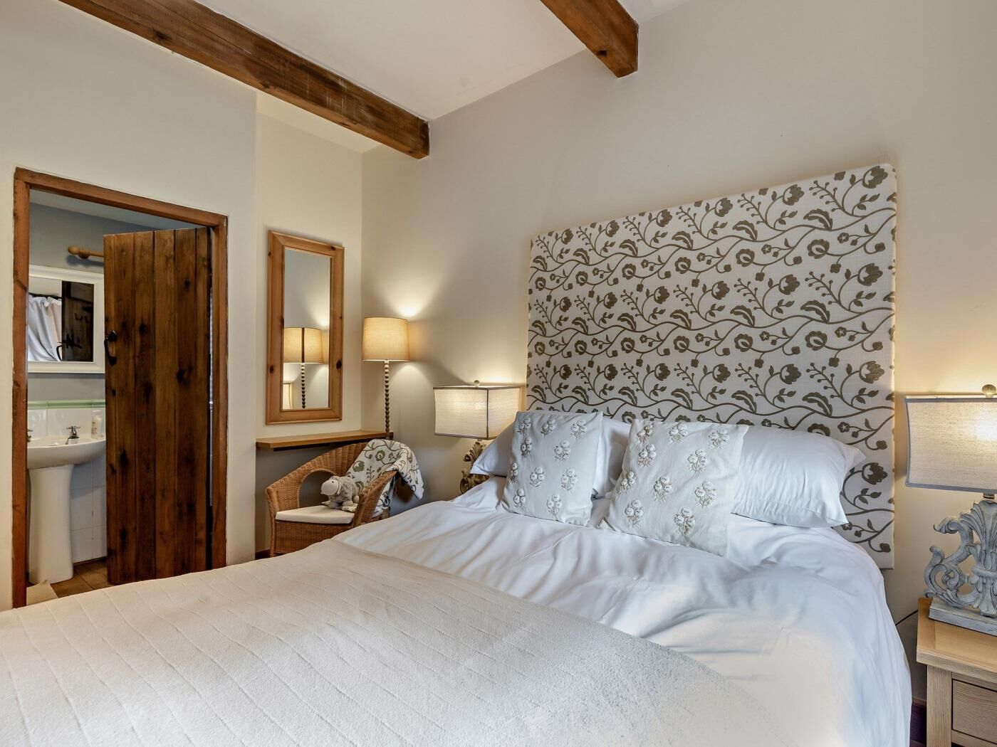 A bedroom with a large bed, a wooden door, a mirror, a bedside table with a lamp, and a patterned headboard.