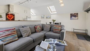 A modern living room with a grey sofa, a coffee table with magazines and a cup, a dining area with a table and chairs, and a kitchen area with a stove and cabinets. - 2 Bed in Beaminster (oc-63047) (Littlewindsor)