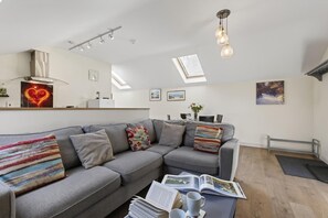 A modern living room with a grey sofa, a coffee table with magazines and a cup, a dining area with a table and chairs, and a kitchen area with a stove and cabinets. - 2 Bed in Beaminster (oc-63047) (Littlewindsor)