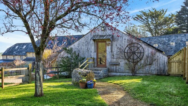 A rustic wooden building with a stone foundation, a wooden door, and a large wheel symbol on the side. - 2 Bed in Beaminster (oc-63047) (Littlewindsor)