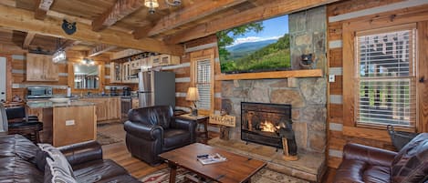 A log cabin interior with a fireplace, leather furniture, a kitchen, and a large window overlooking a landscape.