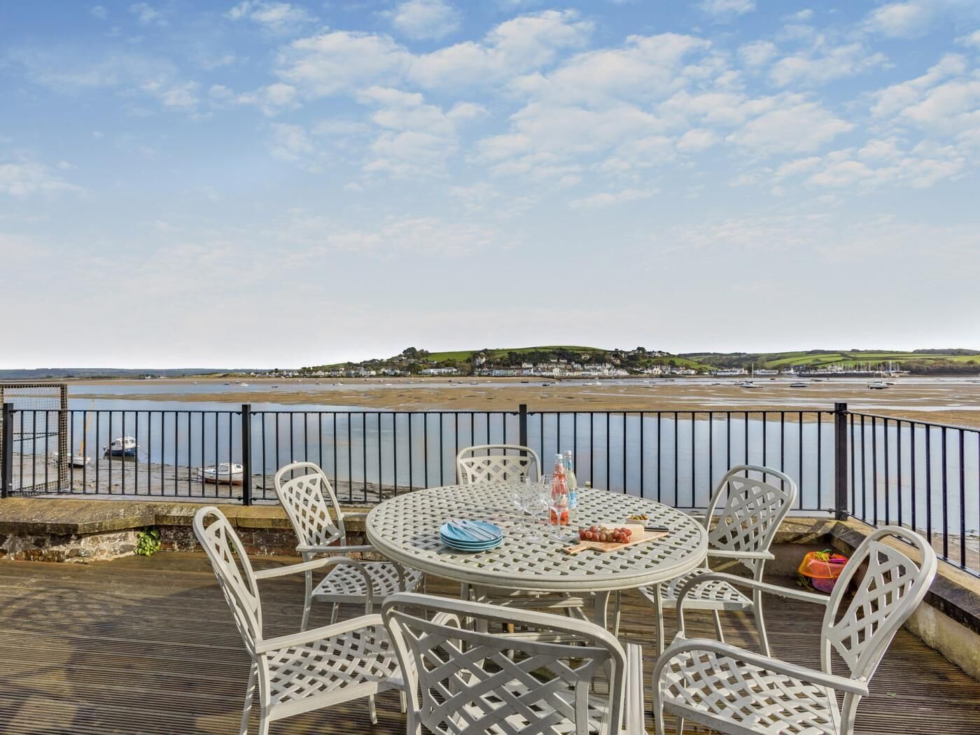 A round table with chairs on a deck overlooking a beach with boats and houses in the distance.