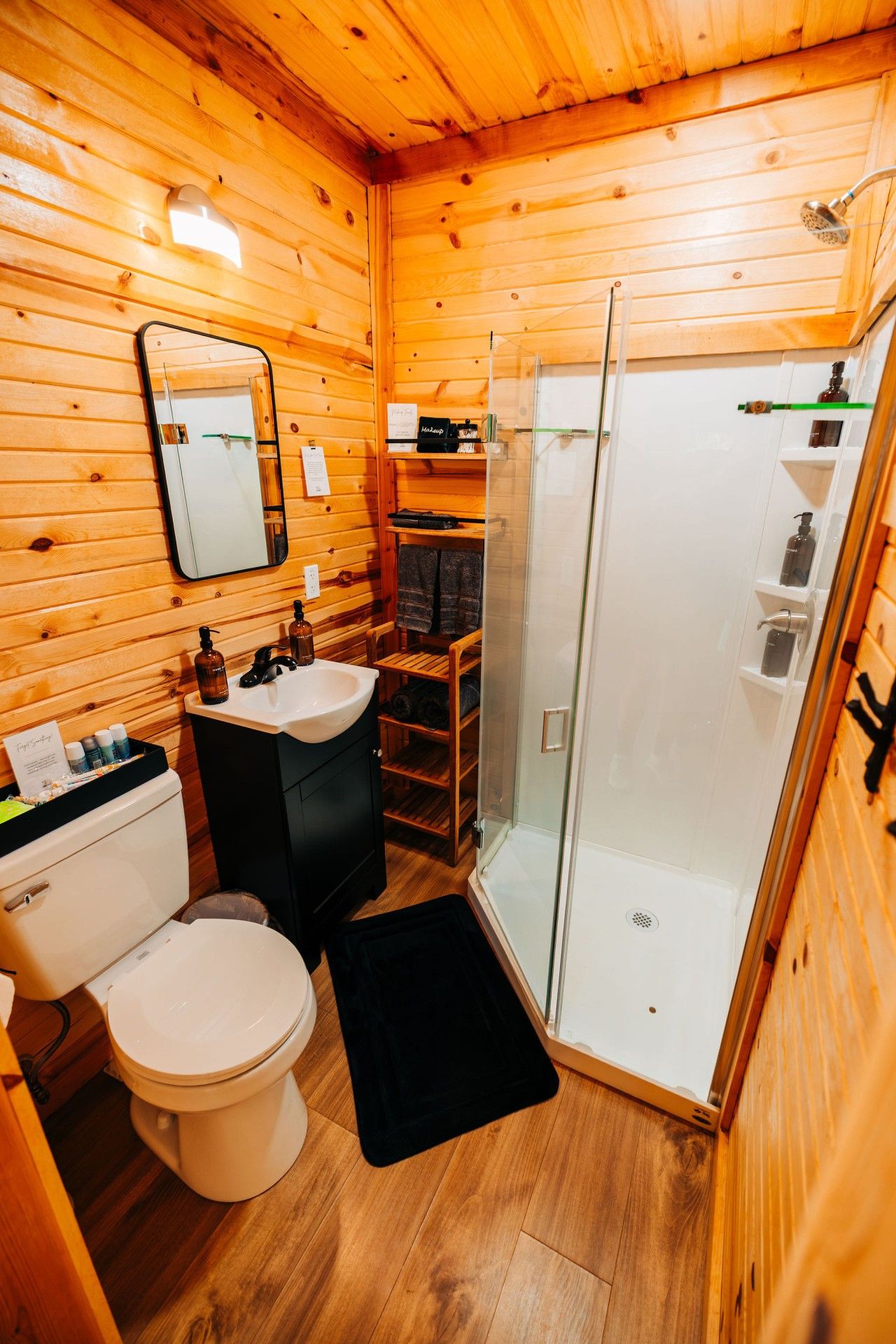 A wooden cabin bathroom with a toilet, sink, and glass shower stall.